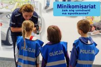 Trójka dzieci przedszkolnych w niebieskich kamizelkach z napisem policja uważnie słucha pochylonej nad nimi policjantki. Policjanta trzymając telefon tłumaczy...