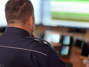 Na zdjęciu oficer dyżurny przed telefonem i ekranem komputera.